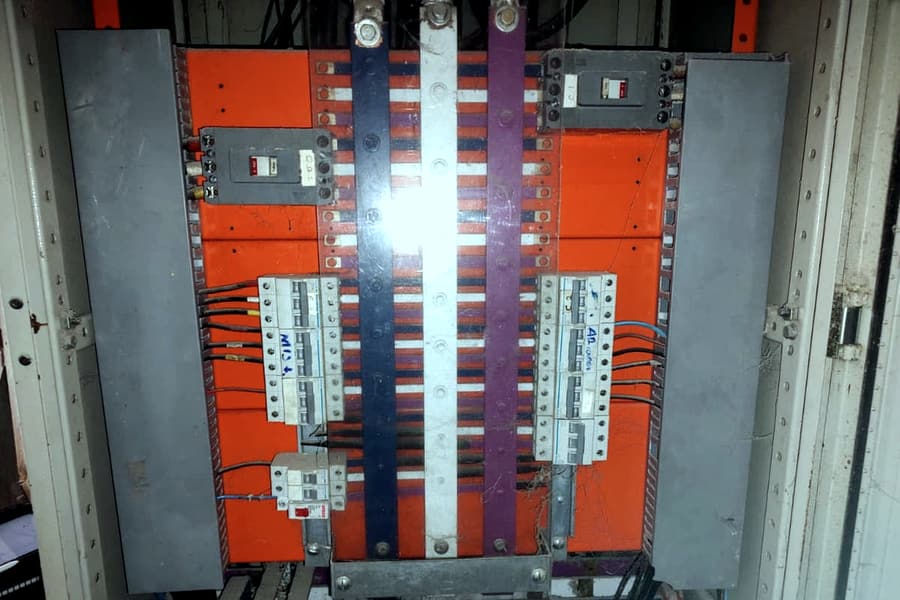 Quadro geral aberto mostrando barramentos horizontais e verticais expostos, com disjuntores laterais e placas de isolação laranja ao fundo.