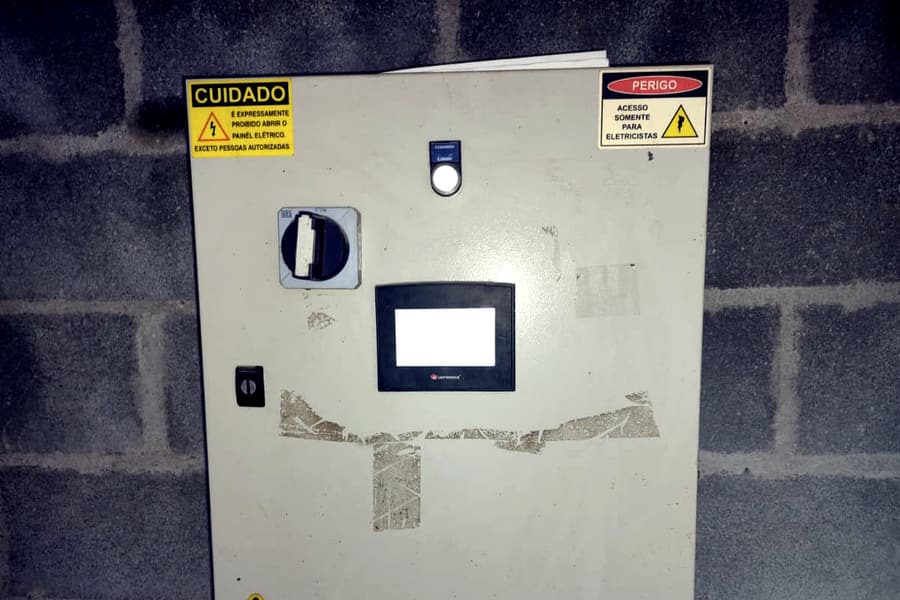 Porta fechada de painel elétrico com placas de advertência, botoeira, chave seccionadora, IHM e restos de adesivos removidos na superfície.